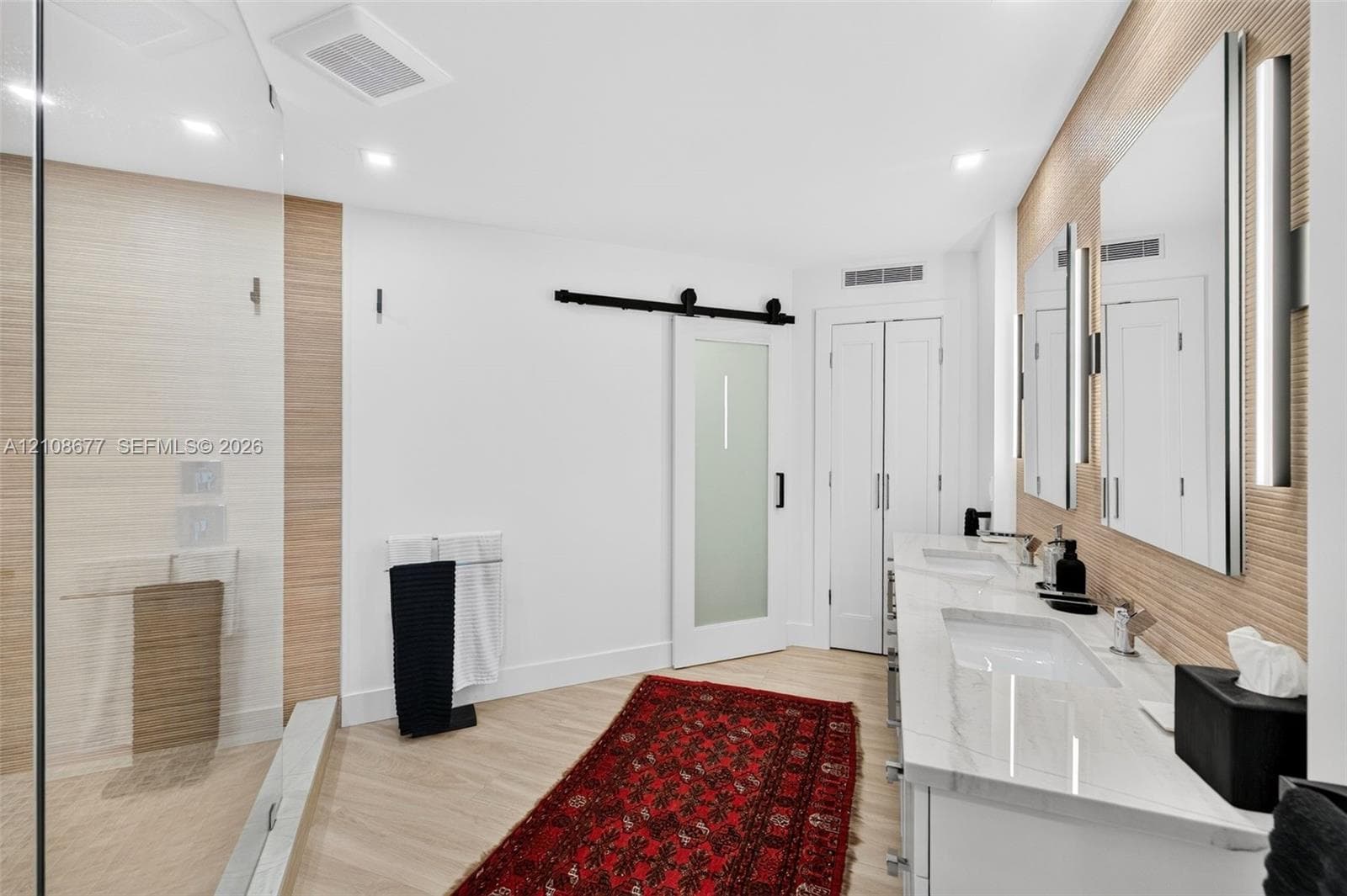 Double vanity with stone-surfaced countertops, frameless glass-enclosed shower, textured wood look wall tile, wall sconces, and frosted glass sliding door to water closet. Tall linen closet next to that.