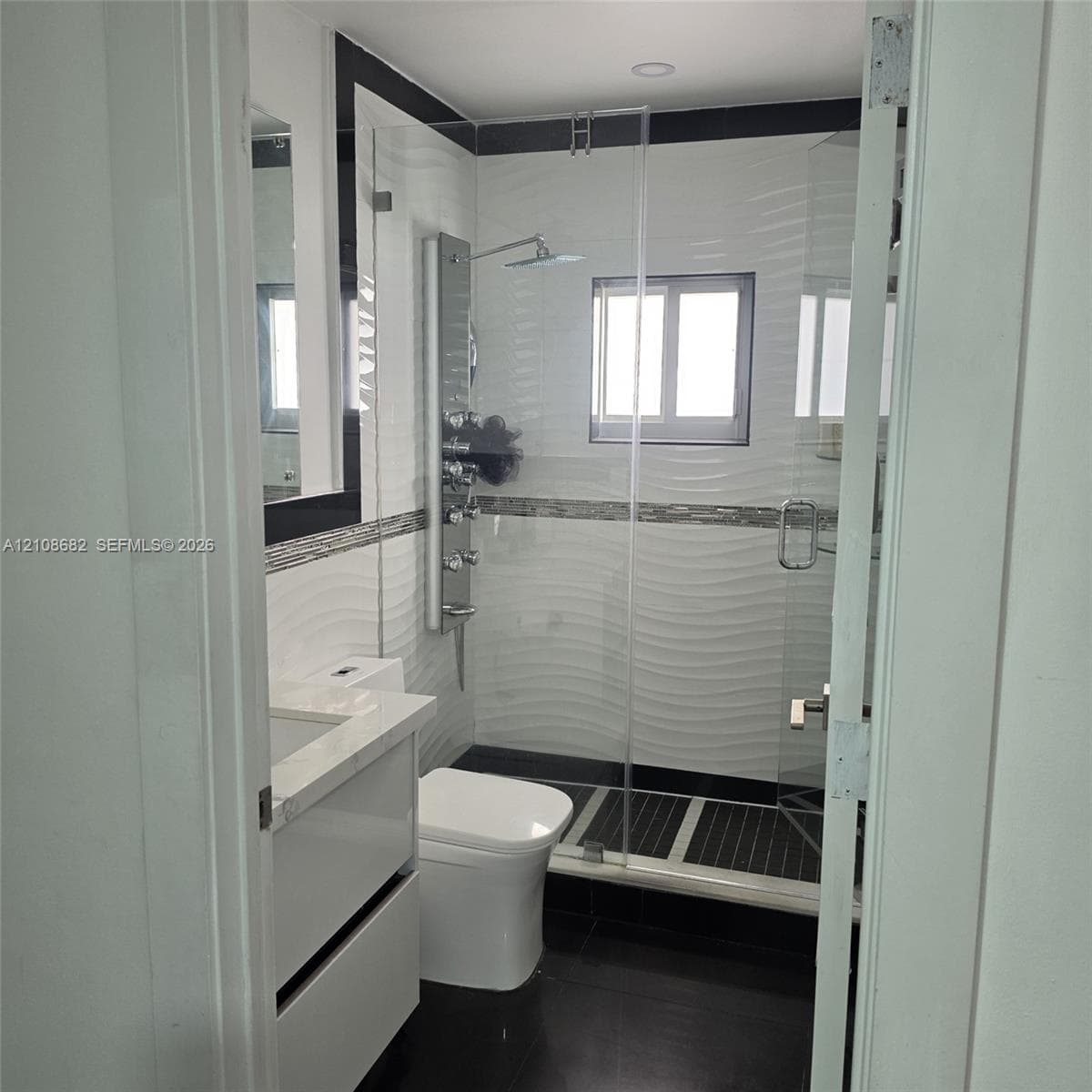 Bathroom featuring a wall-mounted shower panel system, glass shower enclosure, vanity with a stone-top counter, textured tile wall accents, and dark floor tiling