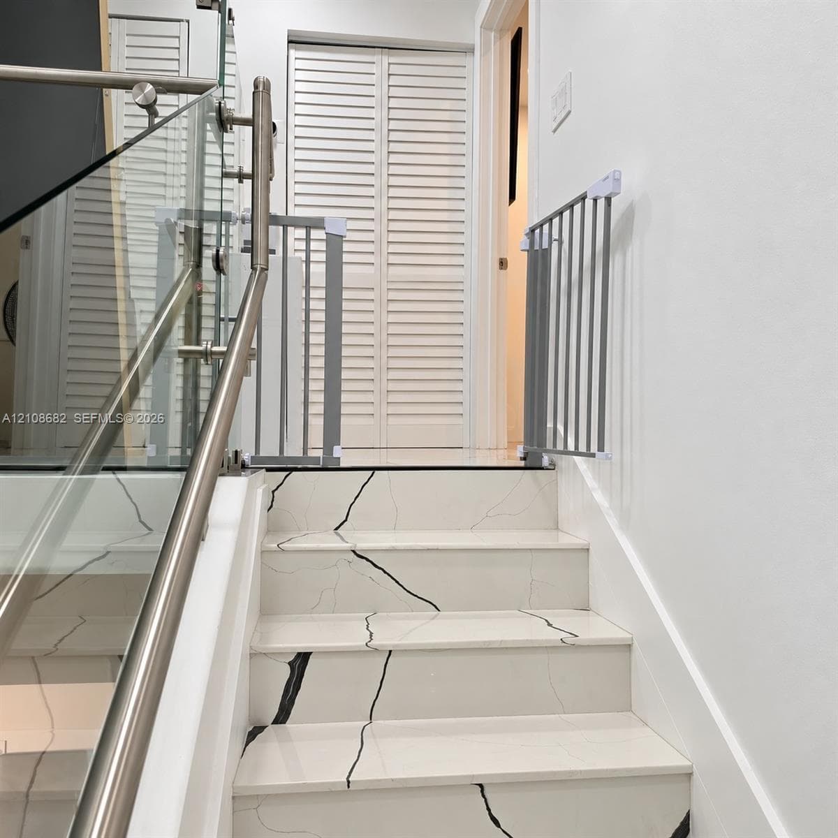 Staircase featuring white stone-finish treads with dark veining, stainless steel handrails, and a glass balustrade, complemented by white louvered closet doors