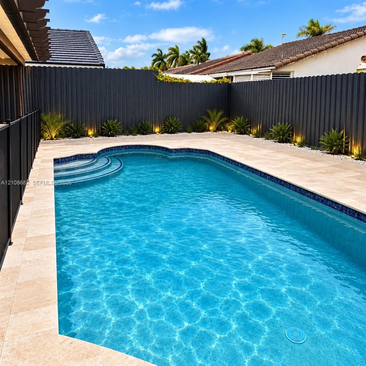 In-ground swimming pool with integrated steps, surrounded by stone paver decking, enclosed by a corrugated metal privacy fence, and featuring perimeter landscape lighting