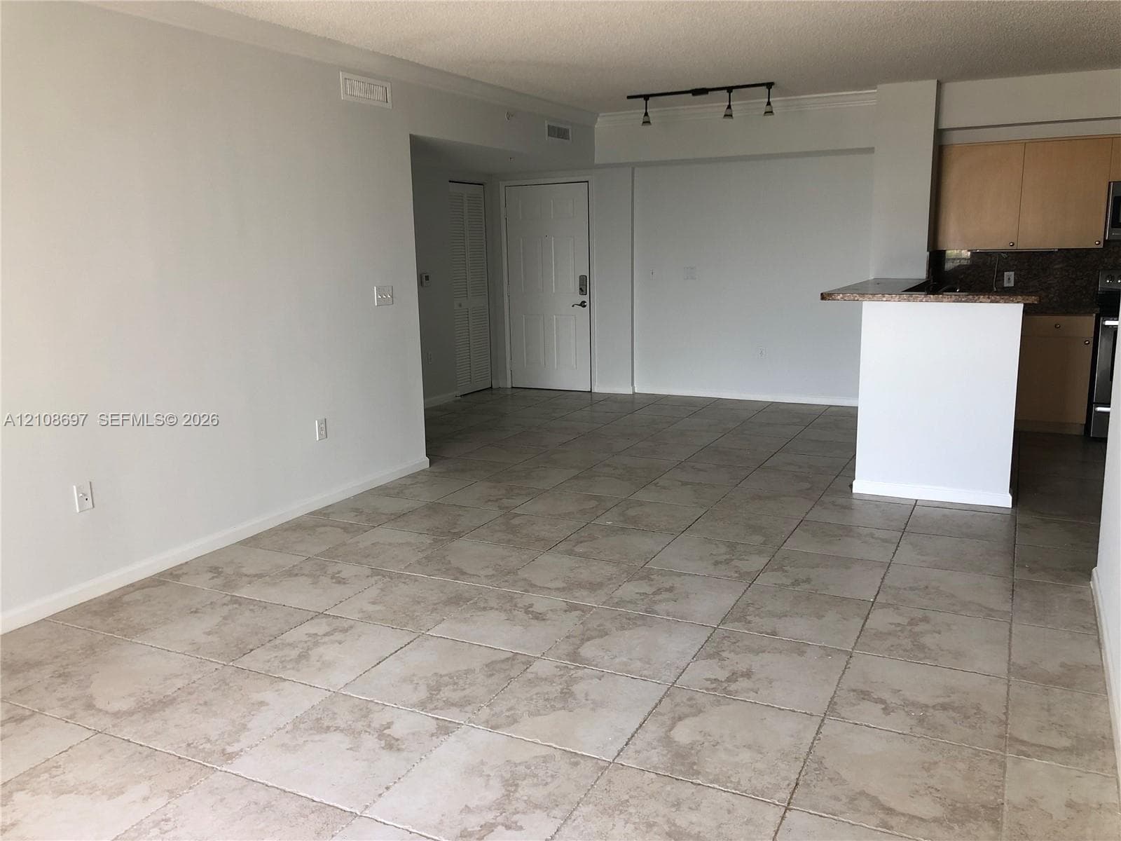 Open living area featuring neutral tile flooring, track lighting, and a kitchen peninsula with a stone-finish countertop and wood cabinetry