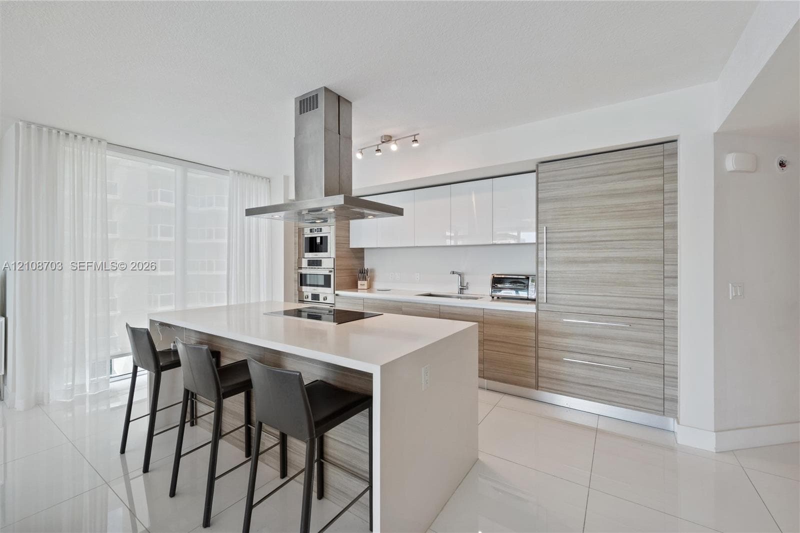 Modern kitchen featuring an island with a waterfall-edge countertop, floor-to-ceiling cabinetry with wood-grain finish, integrated appliances, a stainless steel chimney-style range hood, and large-format tile flooring