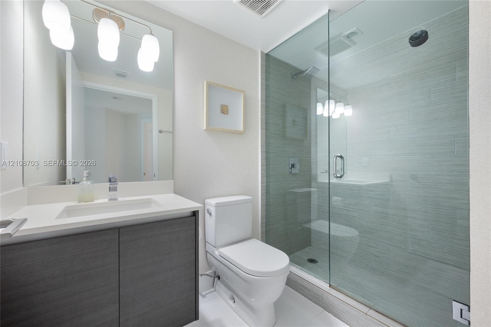 Bathroom featuring a dark wood-finish vanity with a light-toned countertop, a frameless glass-enclosed walk-in shower with textured tile surround, and a wall-mounted light fixture above a large mirror