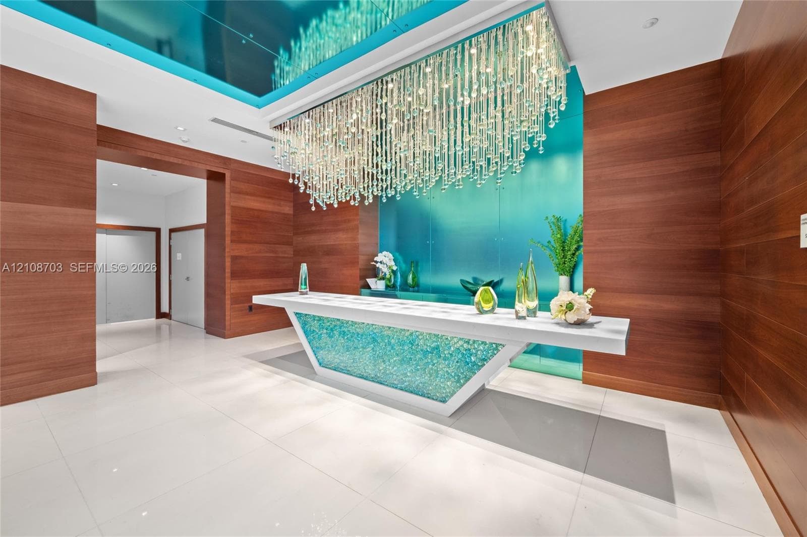 Lobby featuring wood-paneled walls, large-format tile flooring, a geometric reception desk with illuminated glass inlay, and a multi-strand crystal chandelier