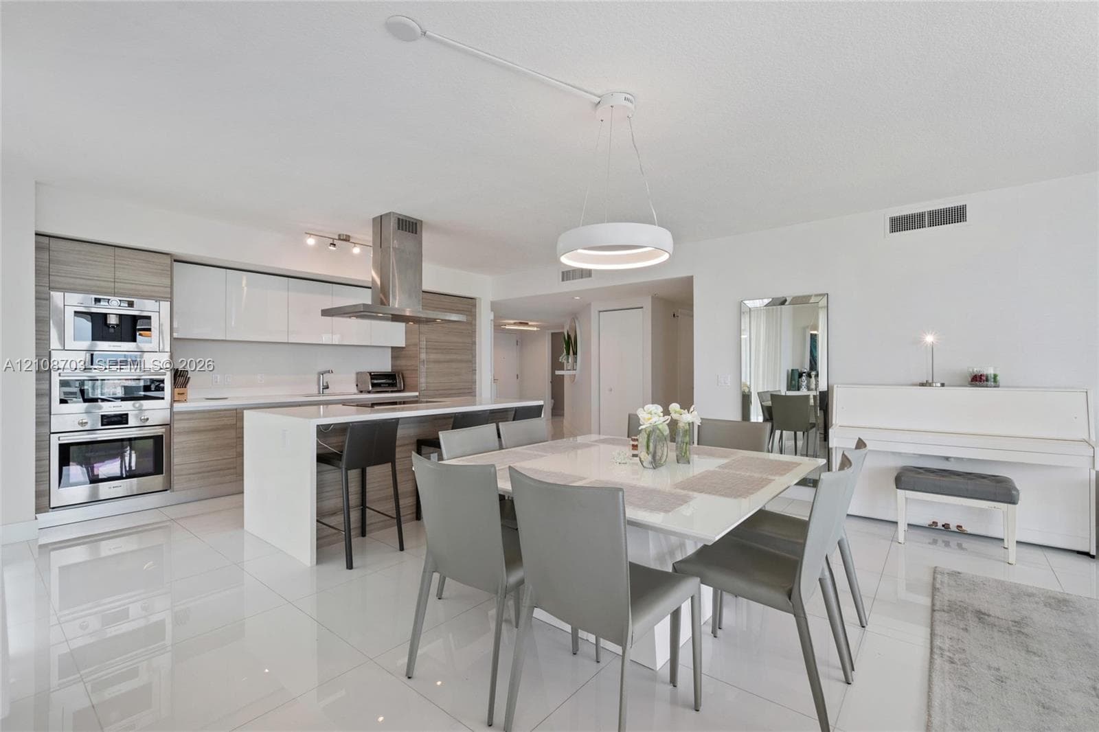 Contemporary kitchen featuring high-gloss cabinetry, stainless steel built-in double ovens, a central island with a waterfall-edge countertop, a stainless steel chimney range hood, and white tile flooring