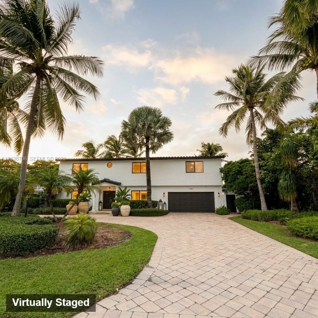 Two-story exterior featuring a light-colored stucco facade, paver driveway, attached multi-car garage, and established tropical landscaping