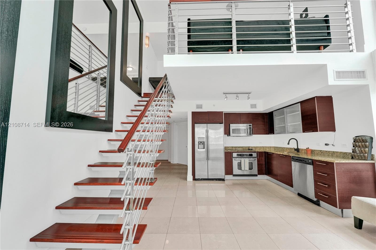 Open-concept interior featuring a stainless steel appliance suite, rich-toned cabinetry with stone-finish countertops, ceramic tile flooring, a mezzanine level with metal railings, and a floating staircase with wood-finish treads and metal cabling