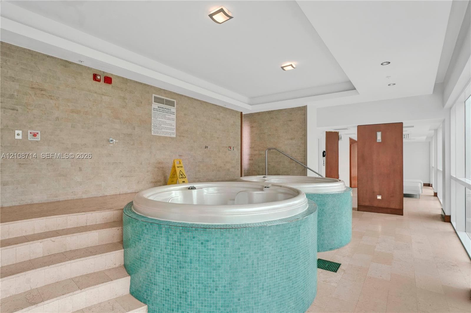 Dual circular jetted tubs with teal mosaic tile surrounds, stone-finish flooring, recessed lighting, and tiered stone stair entry