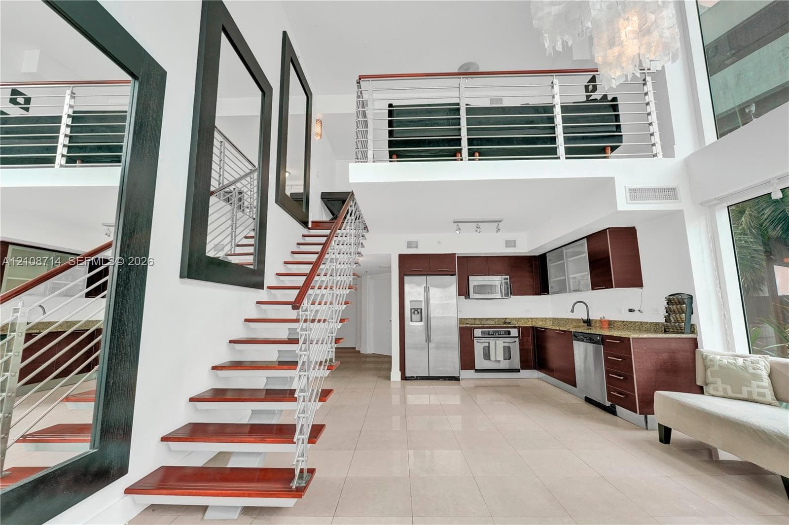 Open-concept interior featuring high ceilings, wood-tread staircase with cable railing, lofted level, tile flooring, and kitchen cabinetry with stainless steel appliances