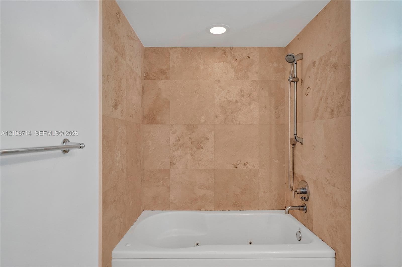 Built-in jetted tub featuring stone-tile surround, adjustable handheld shower fixture, metal faucet, and recessed lighting