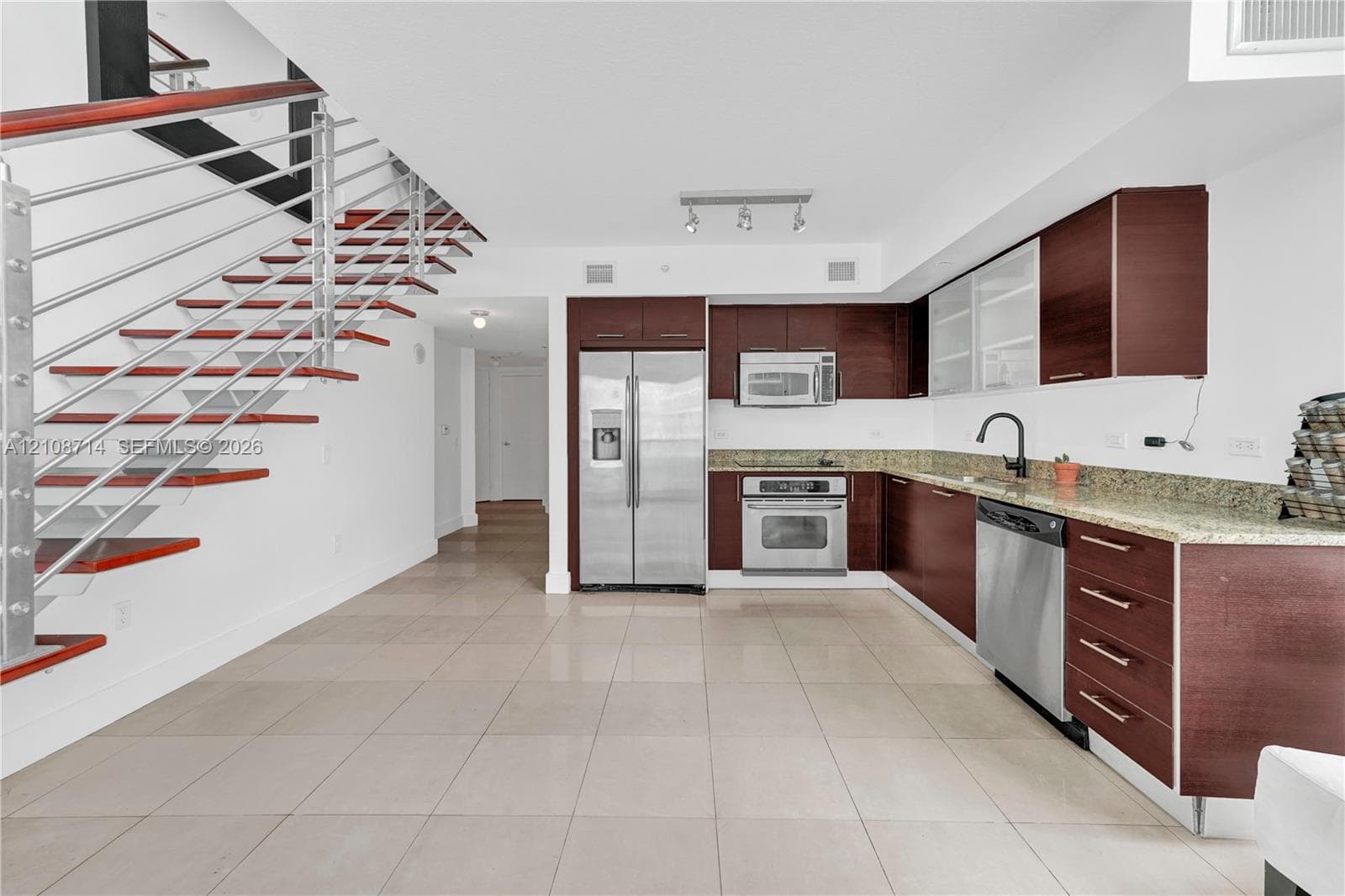 Kitchen featuring wood-finish cabinetry, stone countertops, stainless steel appliances, and large-format tile flooring, accented by an open-riser staircase with a horizontal cable railing system