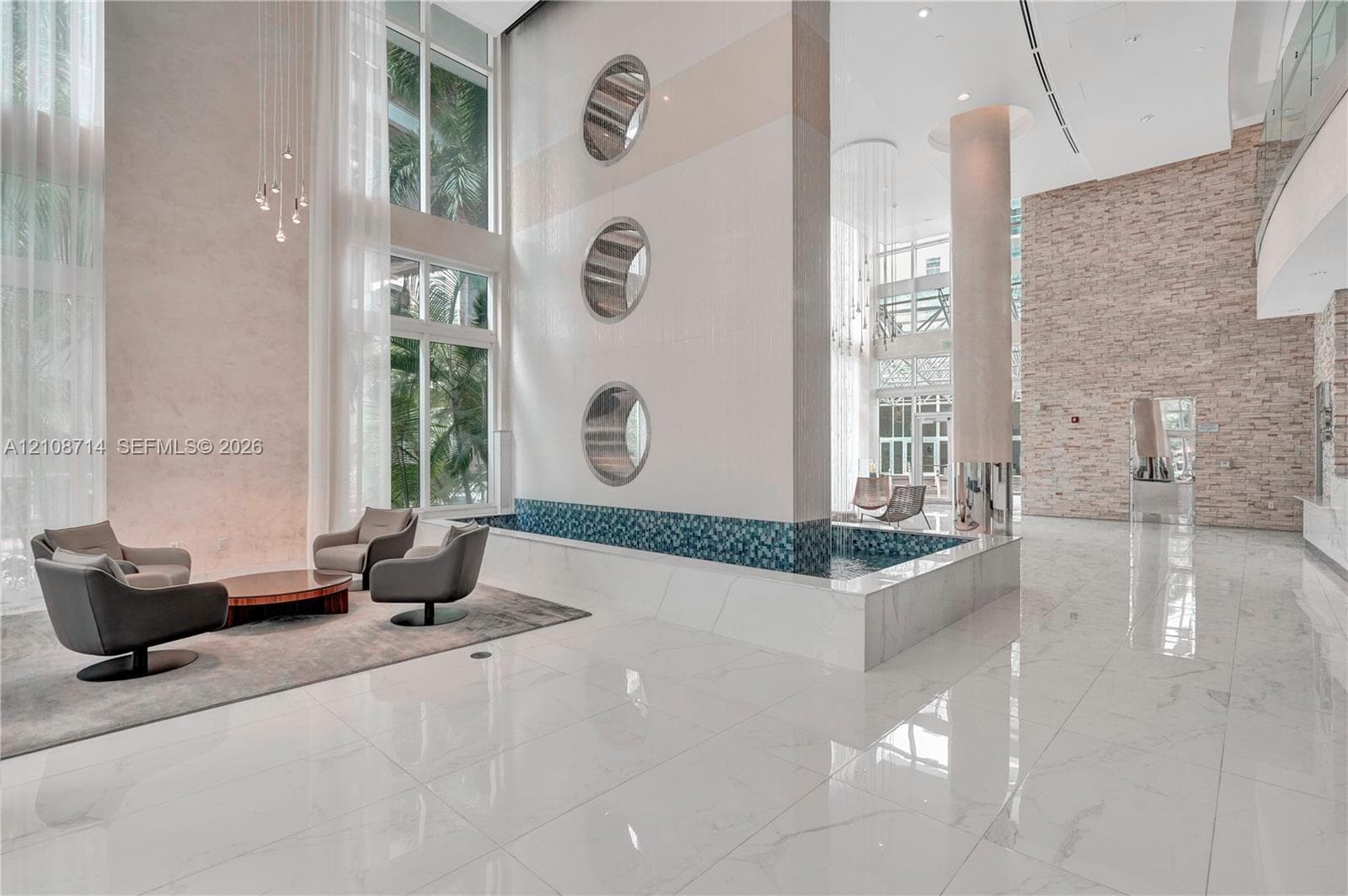 Double-height ceilings featuring floor-to-ceiling windows, polished large-format tile flooring, a decorative indoor water feature with mosaic tile accents, circular wall portals, and textured stone wall cladding