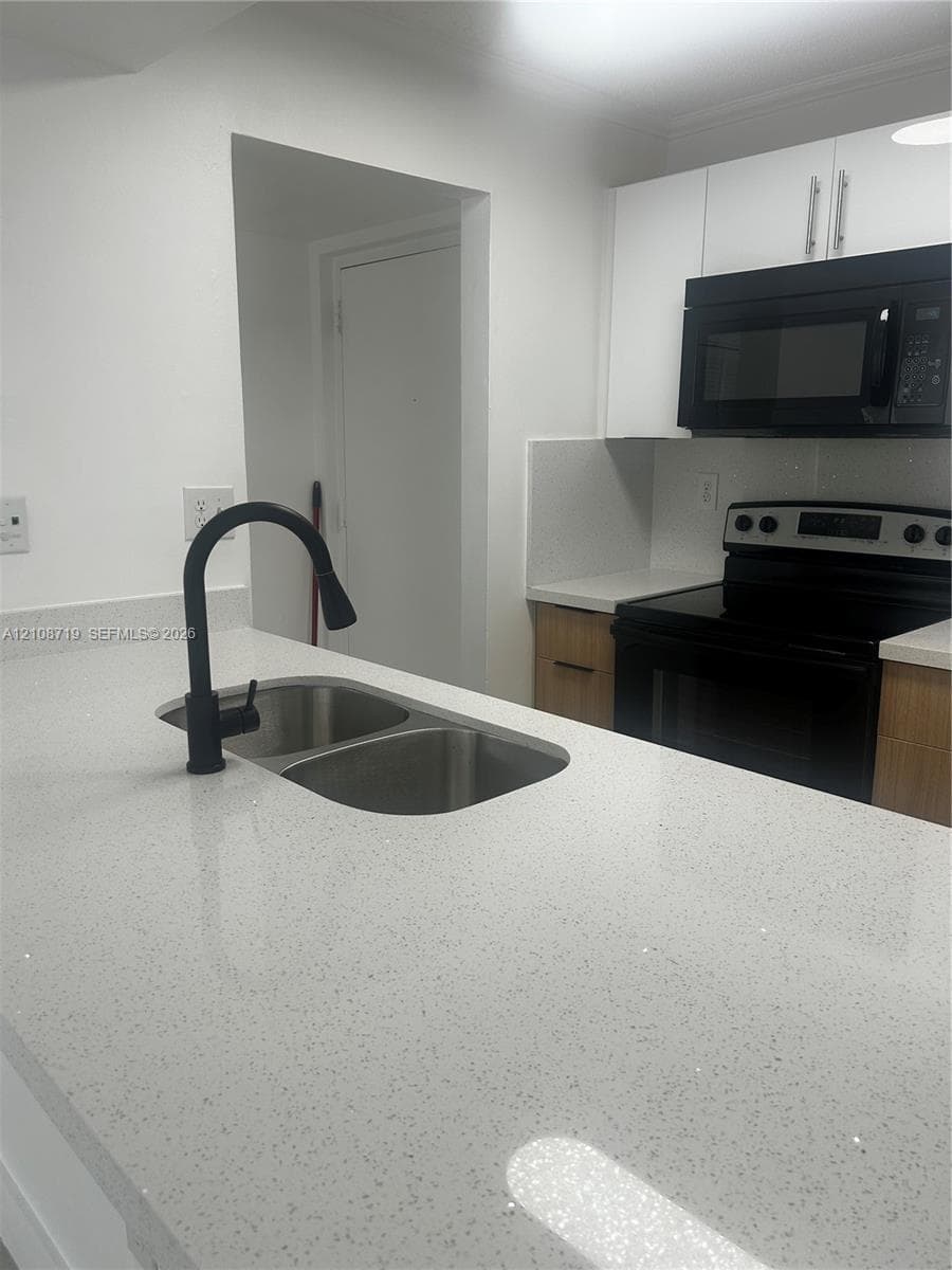 Kitchen featuring a stone-composite countertop, dual-basin stainless steel sink with a matte black pull-down faucet, flat-panel wood-tone cabinetry, and black appliances