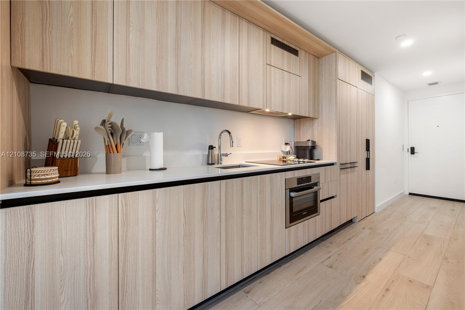 Modern kitchen featuring light wood-finish cabinetry, solid surface countertops, built-in stainless steel appliances, and wide-plank wood-finish flooring