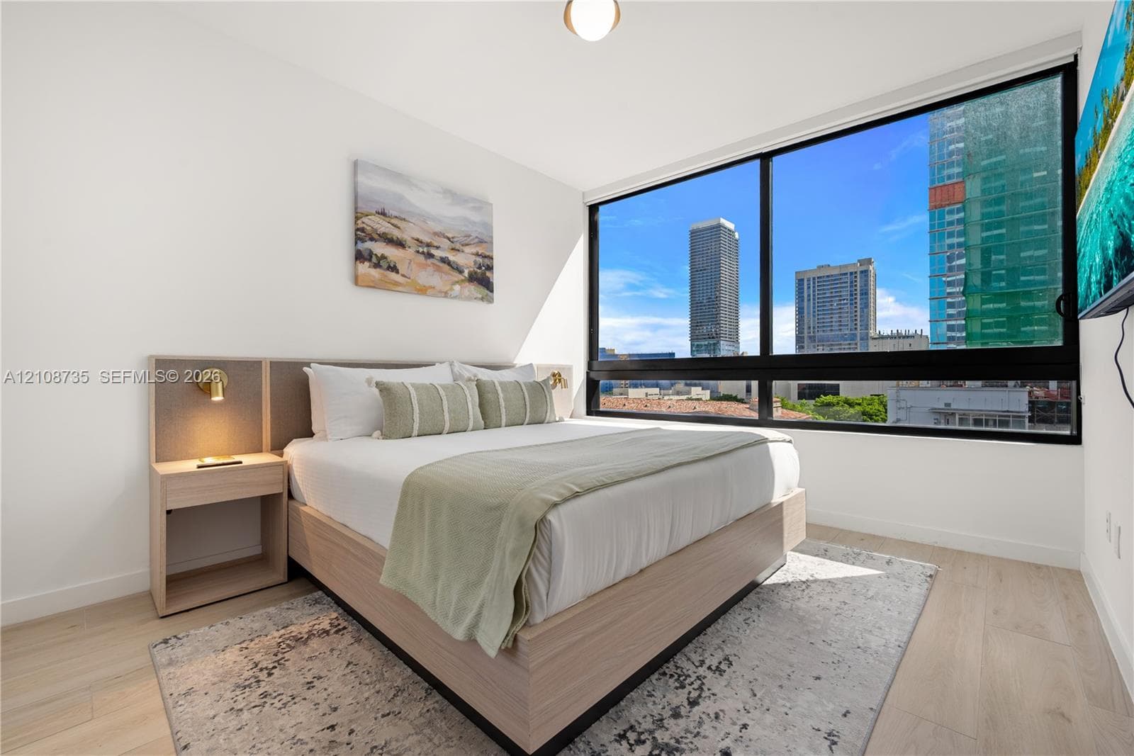 Floor-to-ceiling windows featuring urban scenery, built-in wooden bed frame with integrated headboard, and light wood-finish flooring
