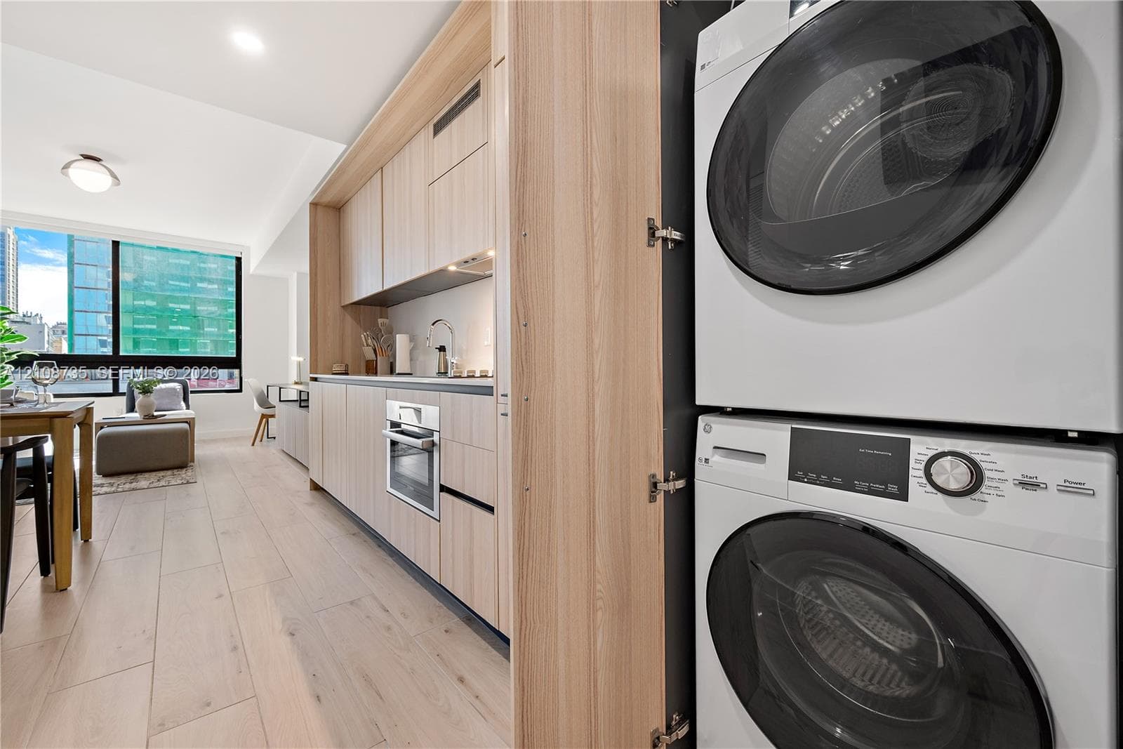 Built-in cabinetry featuring light wood-finish flat-panel doors, integrated stackable laundry appliances, integrated oven, and wood-finish flooring
