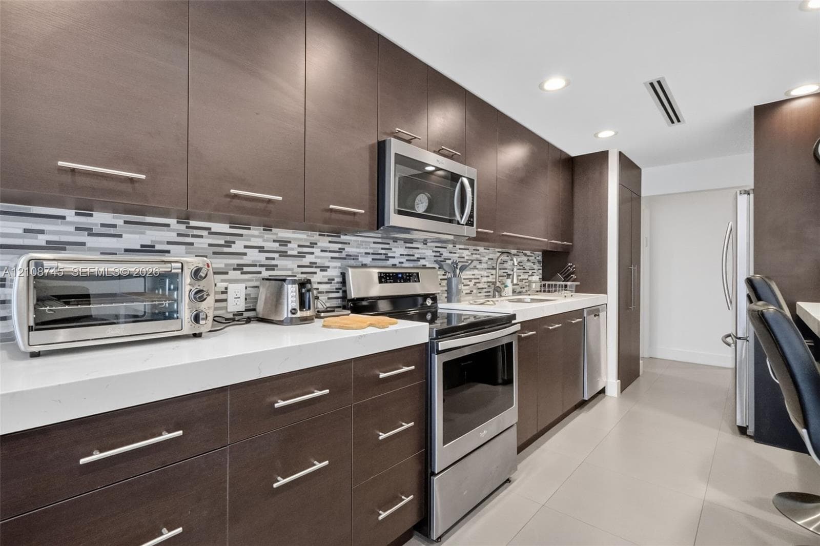 Kitchen featuring dark wood-finish cabinetry, solid surface countertops, a multi-tonal mosaic tile backsplash, and stainless steel appliances