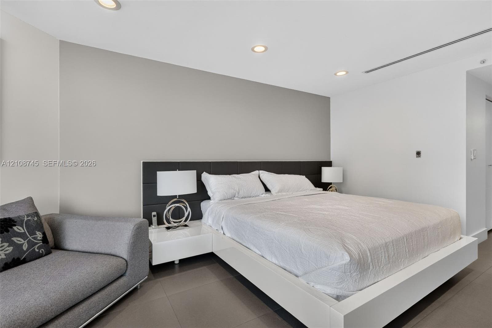 Neutral-toned walls, recessed ceiling lighting, large-format tile flooring, and a built-in dark upholstered headboard