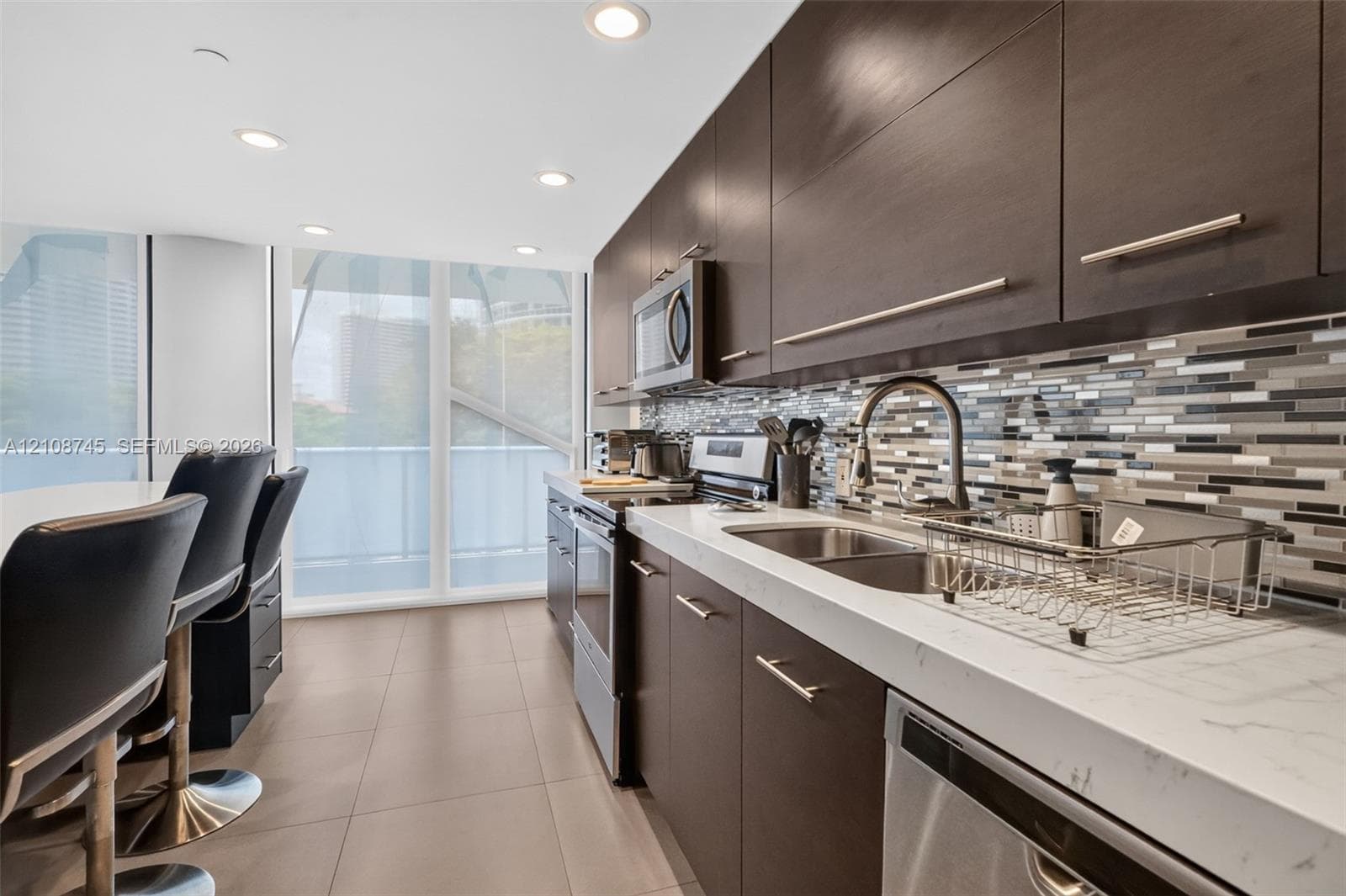 Kitchen featuring dark-toned flat-panel cabinetry, light-colored stone-finish countertops, multi-toned horizontal mosaic tile backsplash, stainless steel appliances, and large-format floor tile