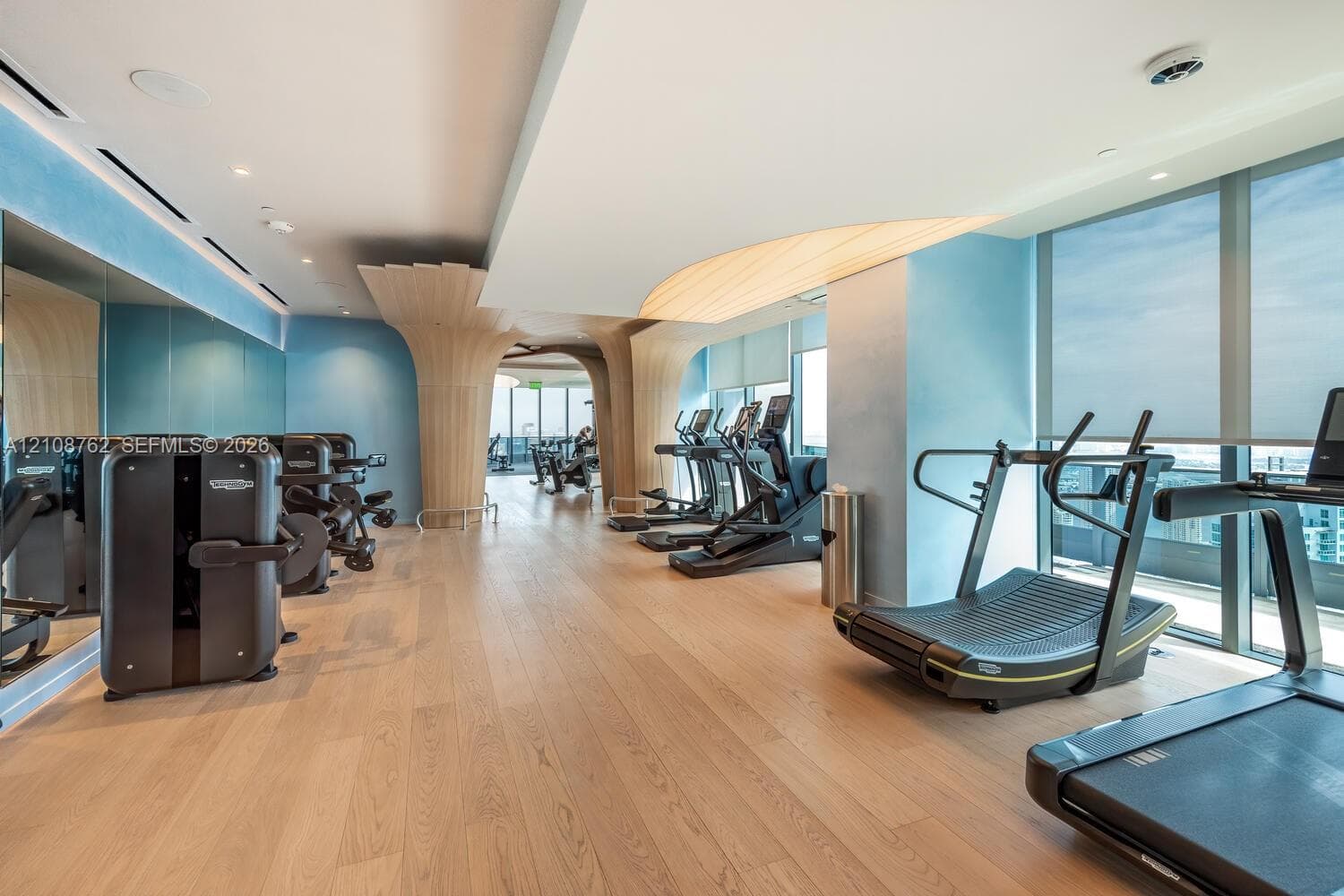 Fitness center featuring wood-finish flooring, floor-to-ceiling windows, custom architectural wood-finish pillars, and integrated ceiling lighting