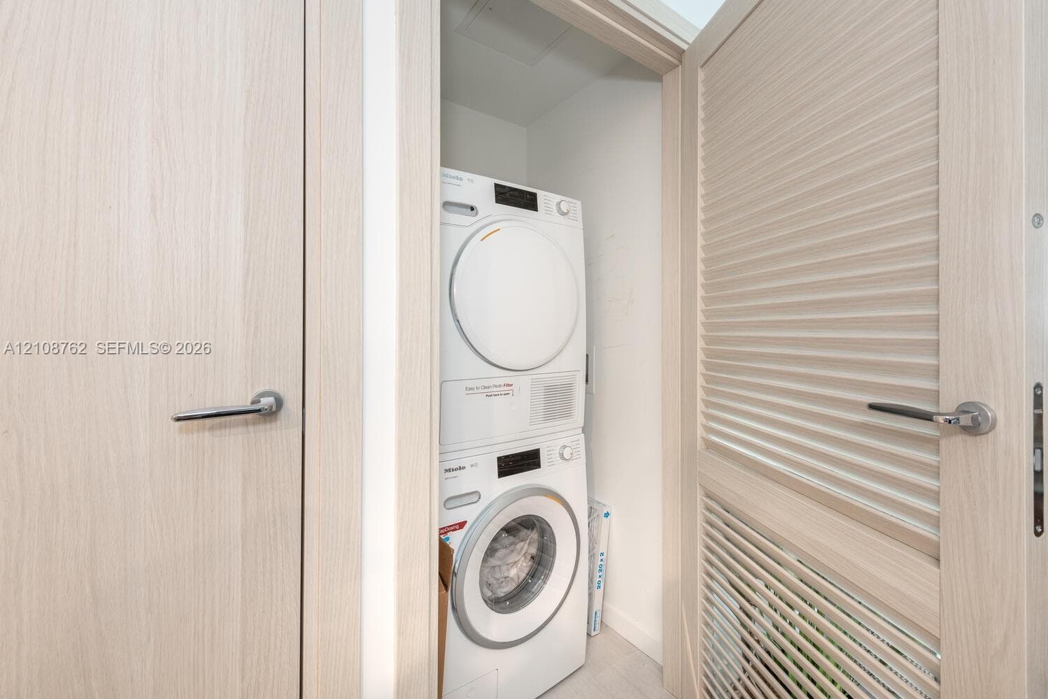 Laundry closet featuring stackable Miele appliances, light-toned wood-finish doors, louvered paneling, and brushed metal hardware