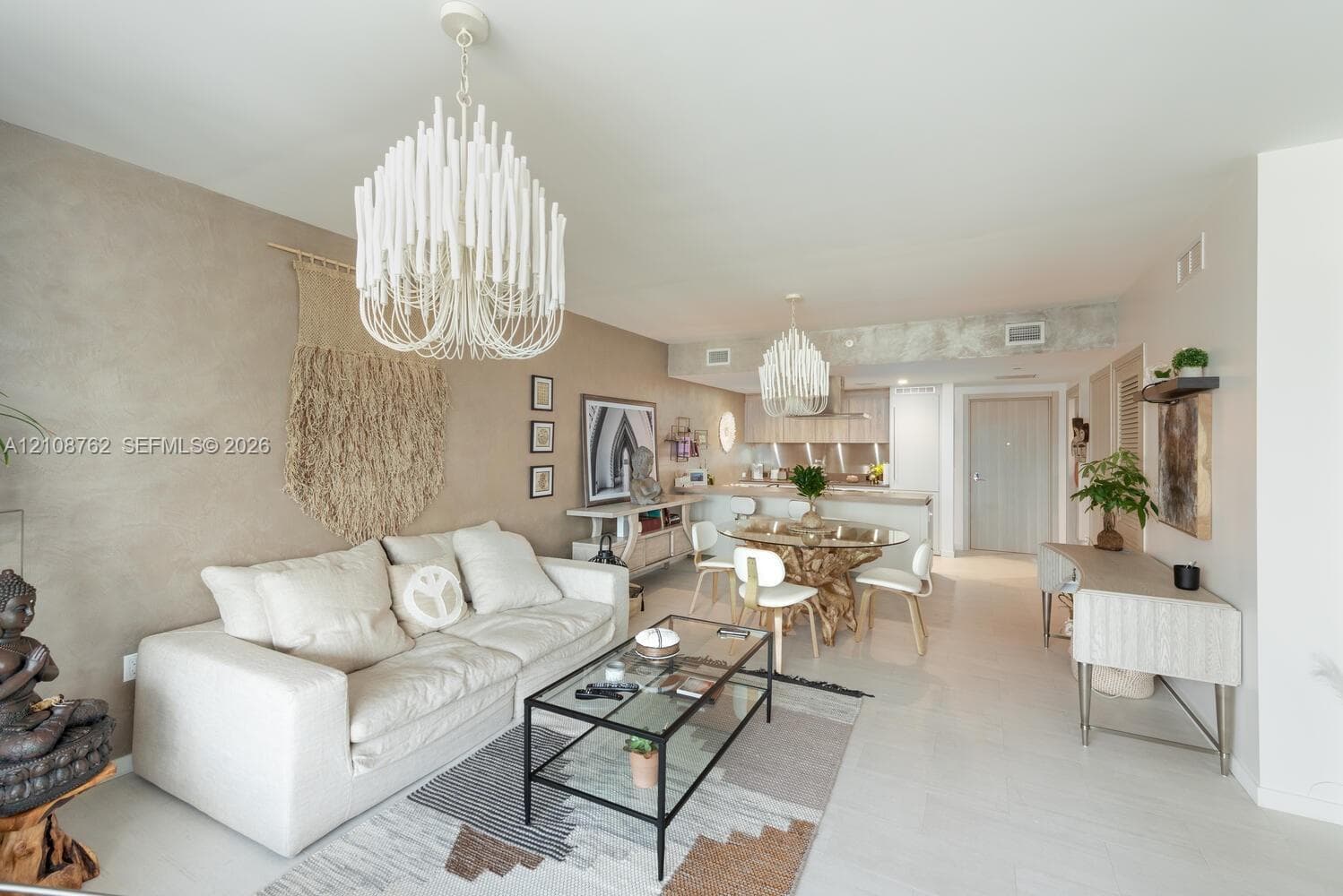 Open-concept living space featuring textured accent walls, light-toned flooring, two tiered candelabra-style chandeliers, and exposed concrete structural elements