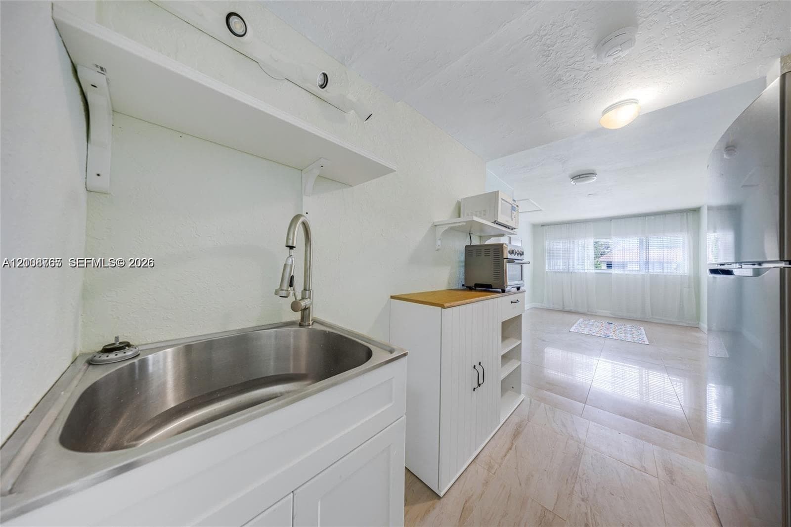 Stainless steel sink with a high-arch pull-down faucet, white cabinetry, light-toned stone-finish flooring, and a stainless steel refrigerator