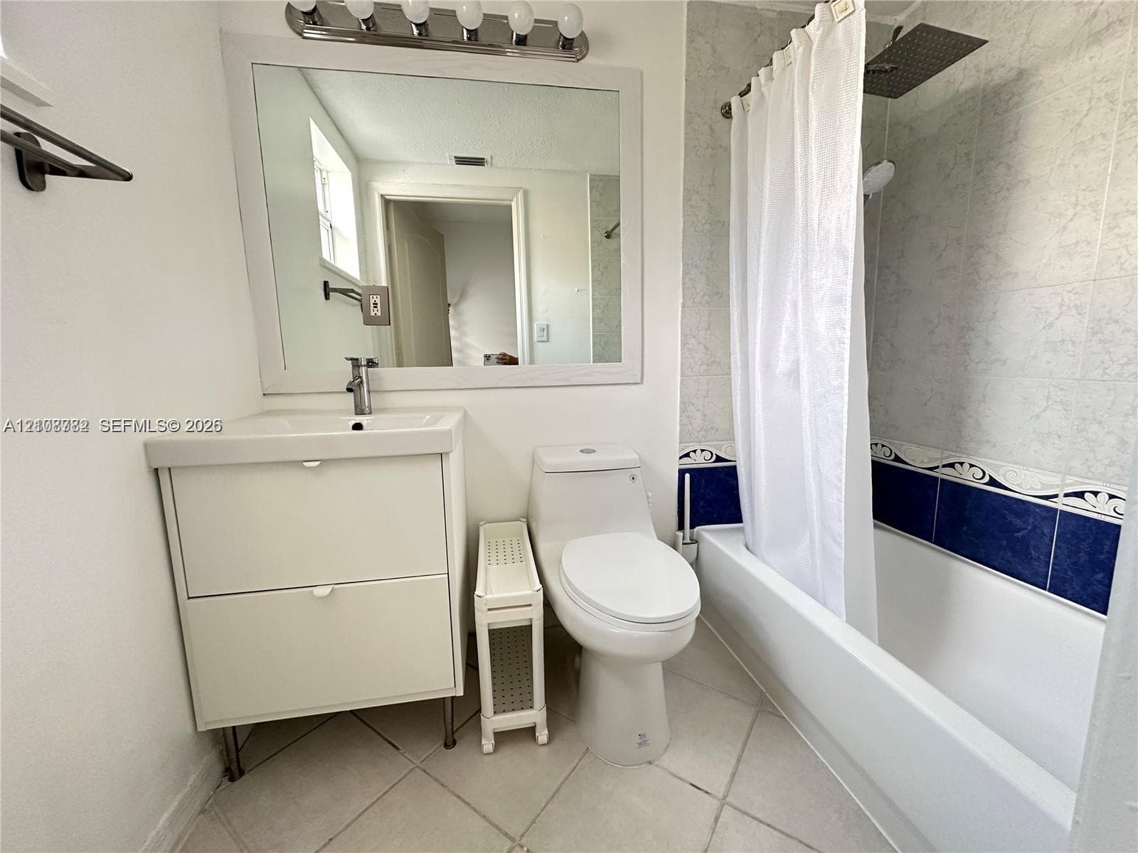 Bathroom featuring a white vanity cabinet with an integrated sink, wall-mounted mirror with integrated light fixture, full-height tile shower surround, and square floor tiling