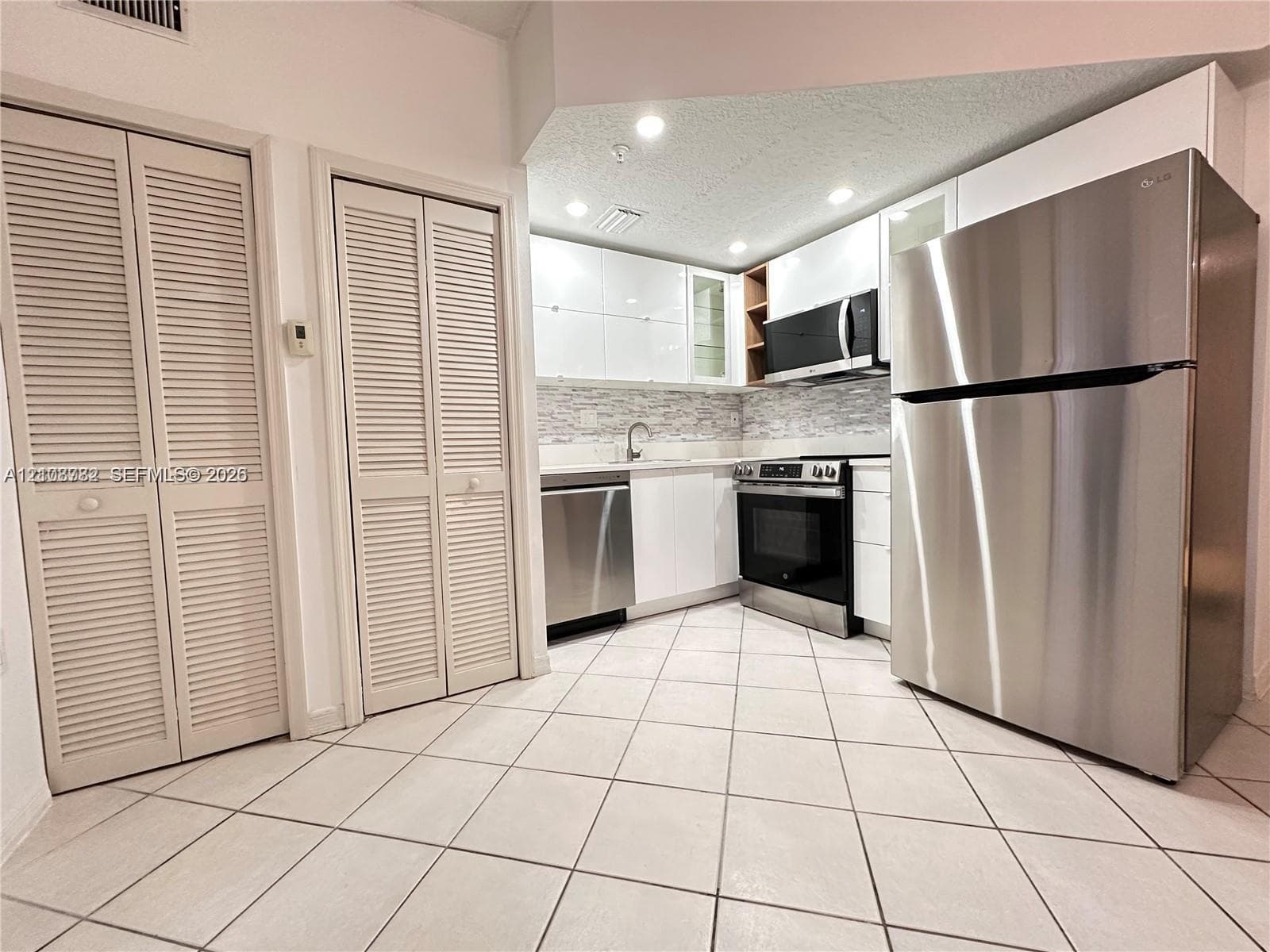 Kitchen featuring white high-gloss cabinetry, stainless steel appliances, a textured stone backsplash, and neutral tile flooring