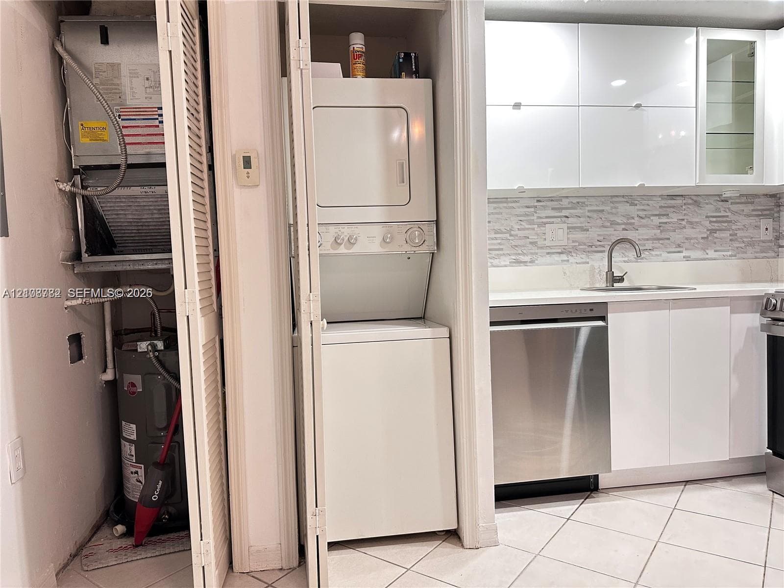 Kitchen featuring high-gloss white cabinetry, stainless steel dishwasher, mosaic tile backsplash, and stacked laundry unit