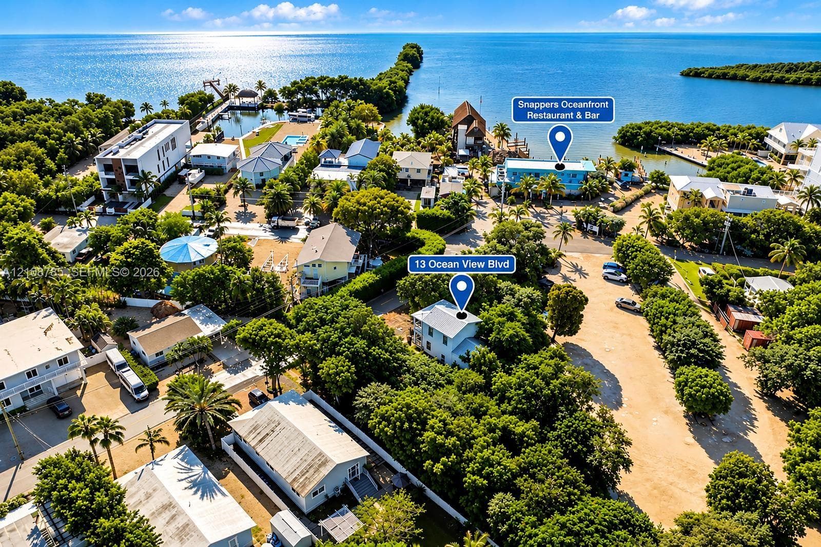Aerial perspective of coastal residential property featuring mature landscaping, metal roofing, ground-level paved parking, and proximity to the open water
