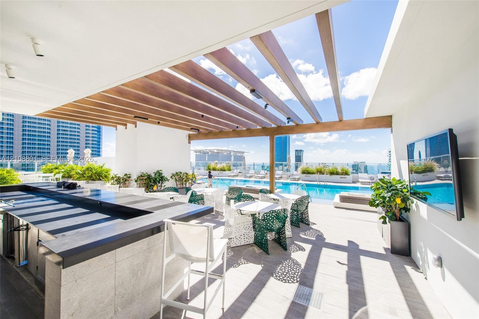 Covered outdoor terrace featuring a wooden pergola, built-in stone-finished bar with integrated cabinetry, porcelain tile flooring, and a mounted flat-screen television, overlooking a swimming pool