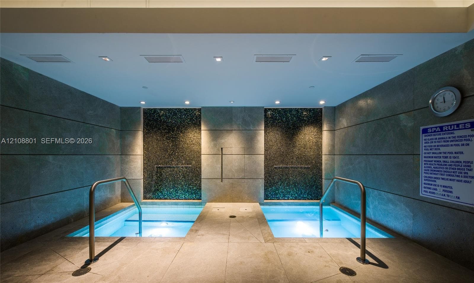 Indoor dual-basin soaking area featuring large-format stone tile wall cladding, mosaic tile accent panels, recessed ceiling lighting, and stainless steel handrails