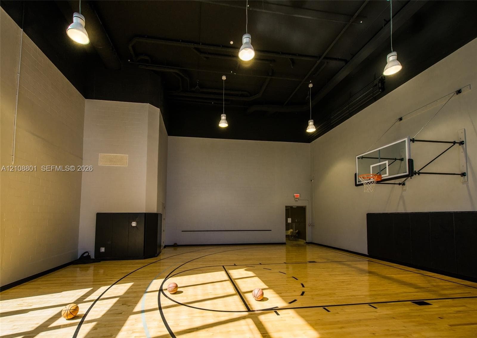 Indoor sports court featuring wood-finish flooring, high ceilings with exposed industrial-style lighting, wall-mounted basketball hoop, and protective wall padding