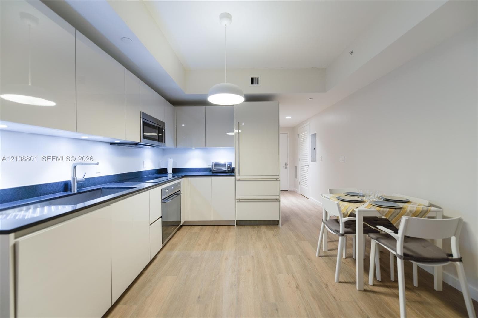 Modern kitchen featuring gloss-finish cabinetry, dark solid-surface countertops, integrated stainless steel appliances, under-cabinet lighting, and wood-finish flooring