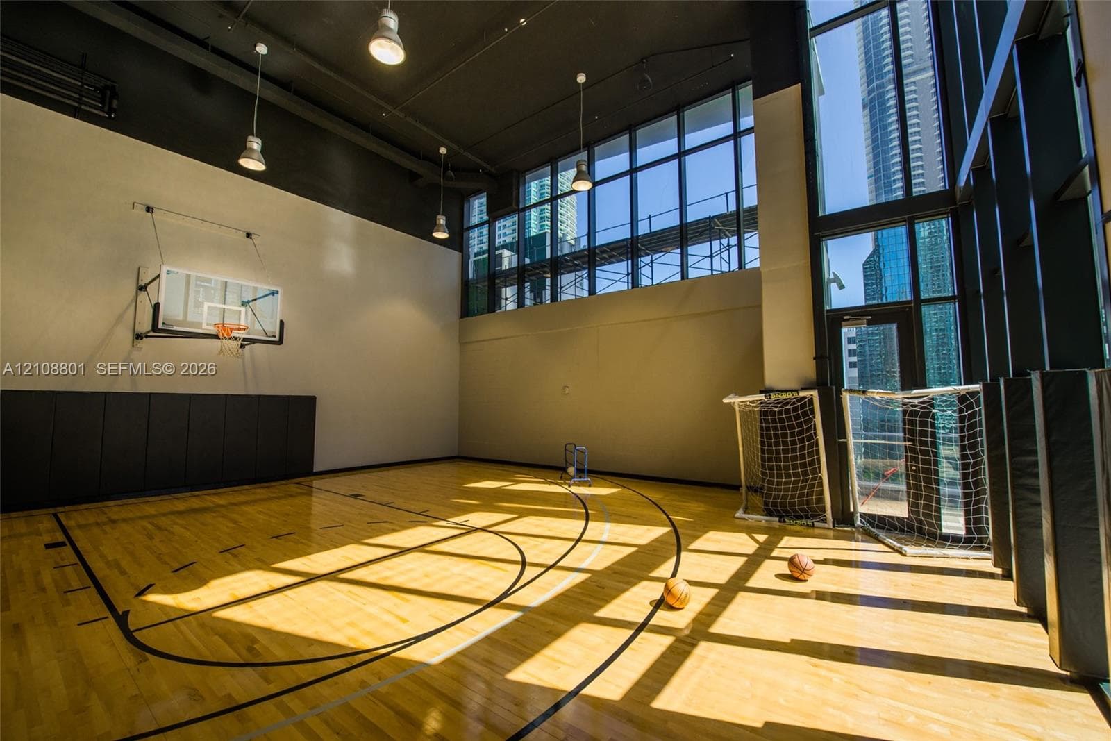 Indoor sport court featuring wood-finish flooring, high ceilings with pendant lighting, floor-to-ceiling windows, and wall-mounted protective padding