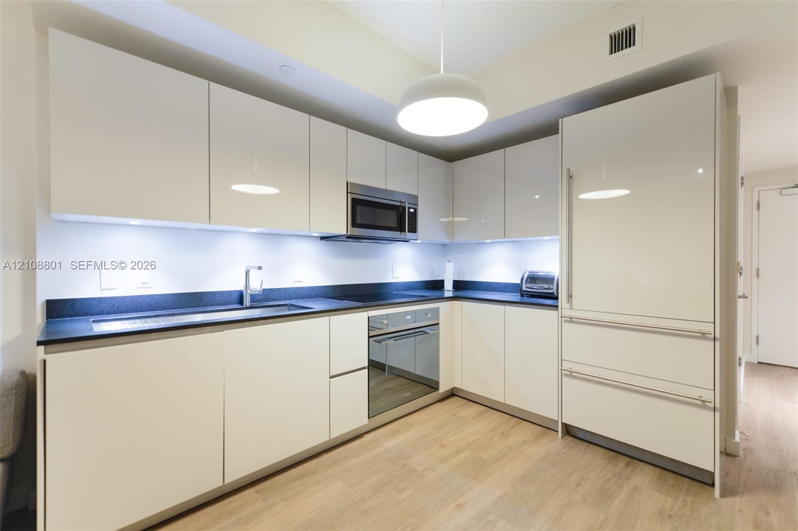 Kitchen featuring high-gloss cabinetry, dark stone-finish countertops, integrated stainless steel appliances, under-cabinet task lighting, and wood-finish flooring