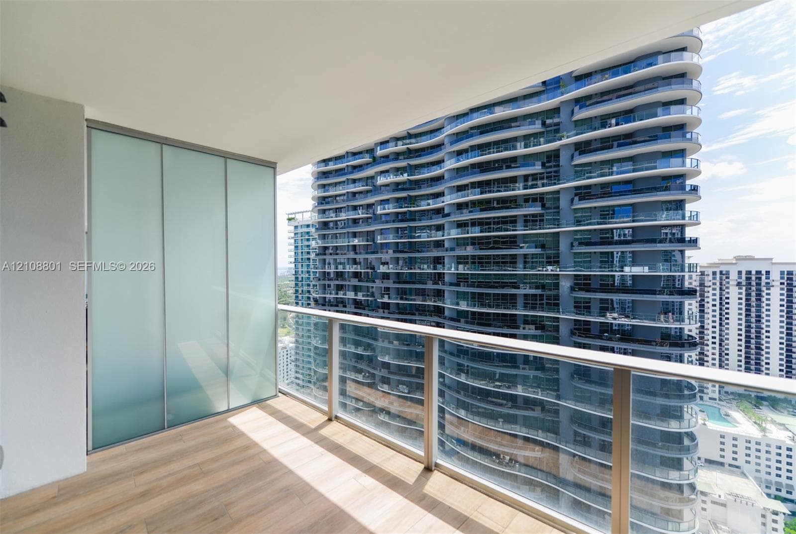 Covered private balcony featuring wood-finish flooring, frosted glass privacy panel, and a structural metal and glass railing