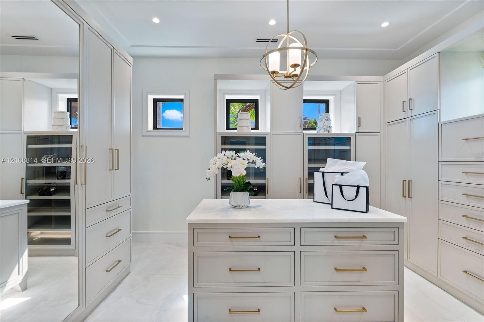 Custom cabinetry featuring glass-front display doors and integrated shelving, central storage island with stone-finish countertop, brushed brass hardware, mirrored door panels, and spherical pendant lighting