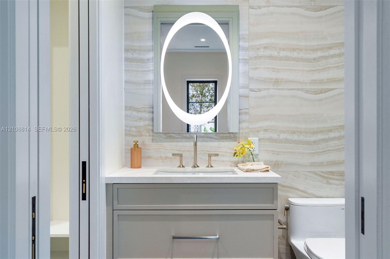 Custom vanity with stone-surface countertop, integrated rectangular sink, oval backlit mirror, and floor-to-ceiling stone-tiled wall