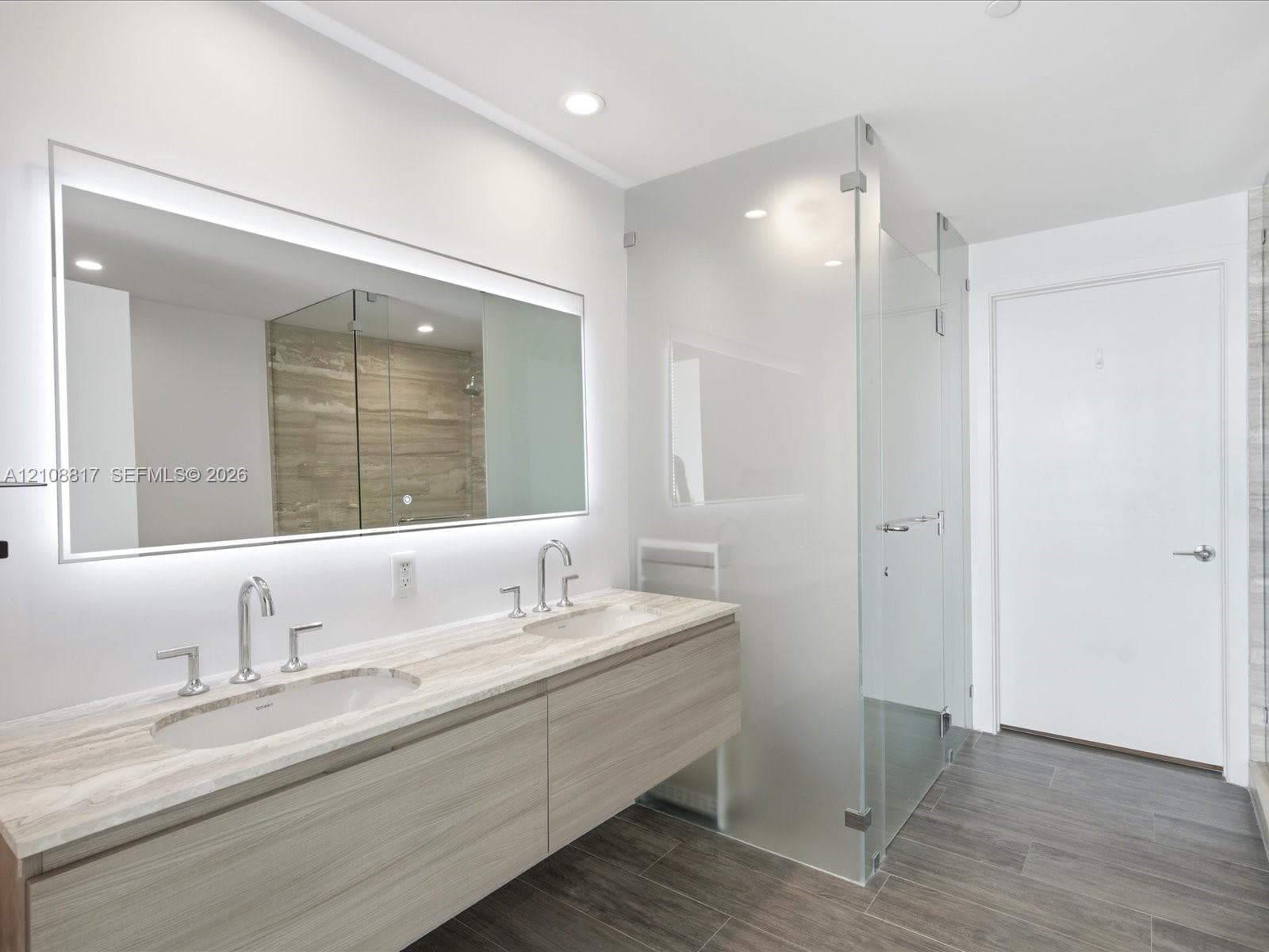 Floating dual-sink vanity featuring stone countertops and polished chrome widespread faucets, complemented by a large back-lit mirror, glass-enclosed shower, and wood-finish tile flooring