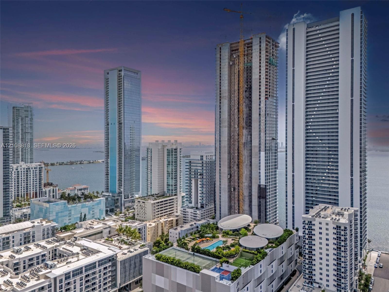 High-rise urban landscape featuring a rooftop terrace with a swimming pool, tennis court, and lush landscaping, set against a coastal backdrop