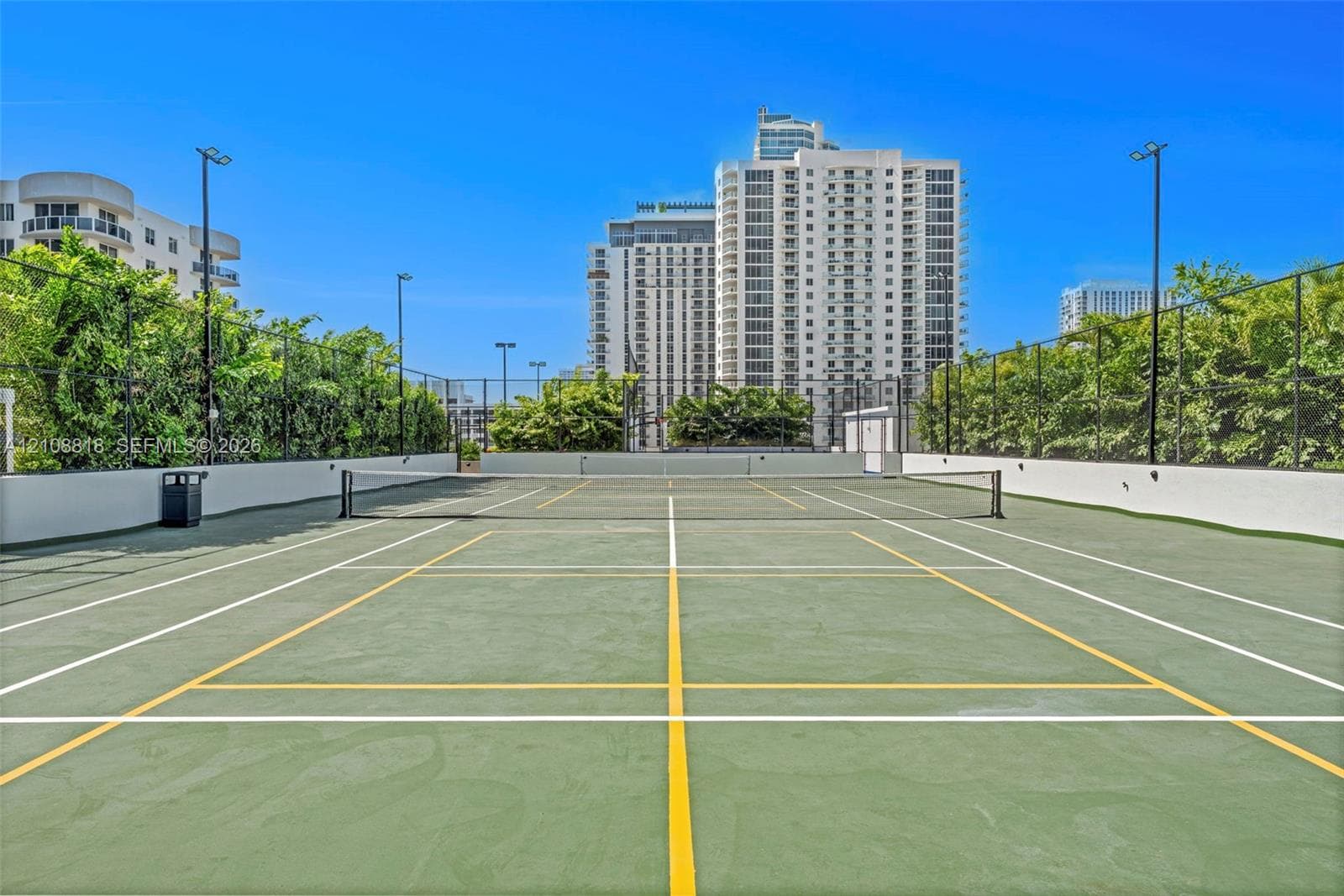 Hard-surfaced tennis court featuring perimeter chain-link fencing, high-mast lighting, and surrounding mature greenery