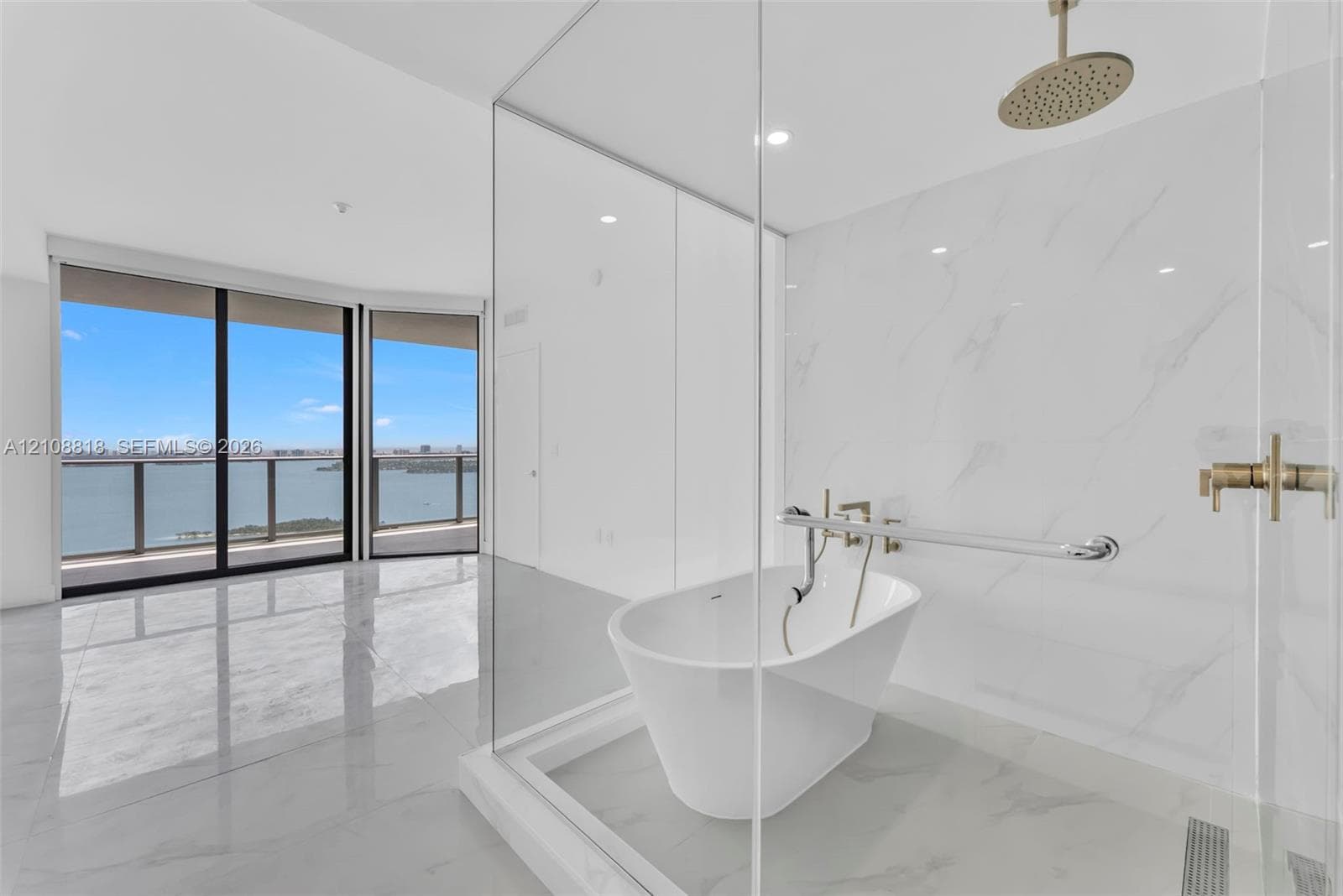 Freestanding soaking tub, glass-enclosed shower area with wall-mounted rainfall showerhead, polished stone-finish flooring, and floor-to-ceiling windows with water backdrop