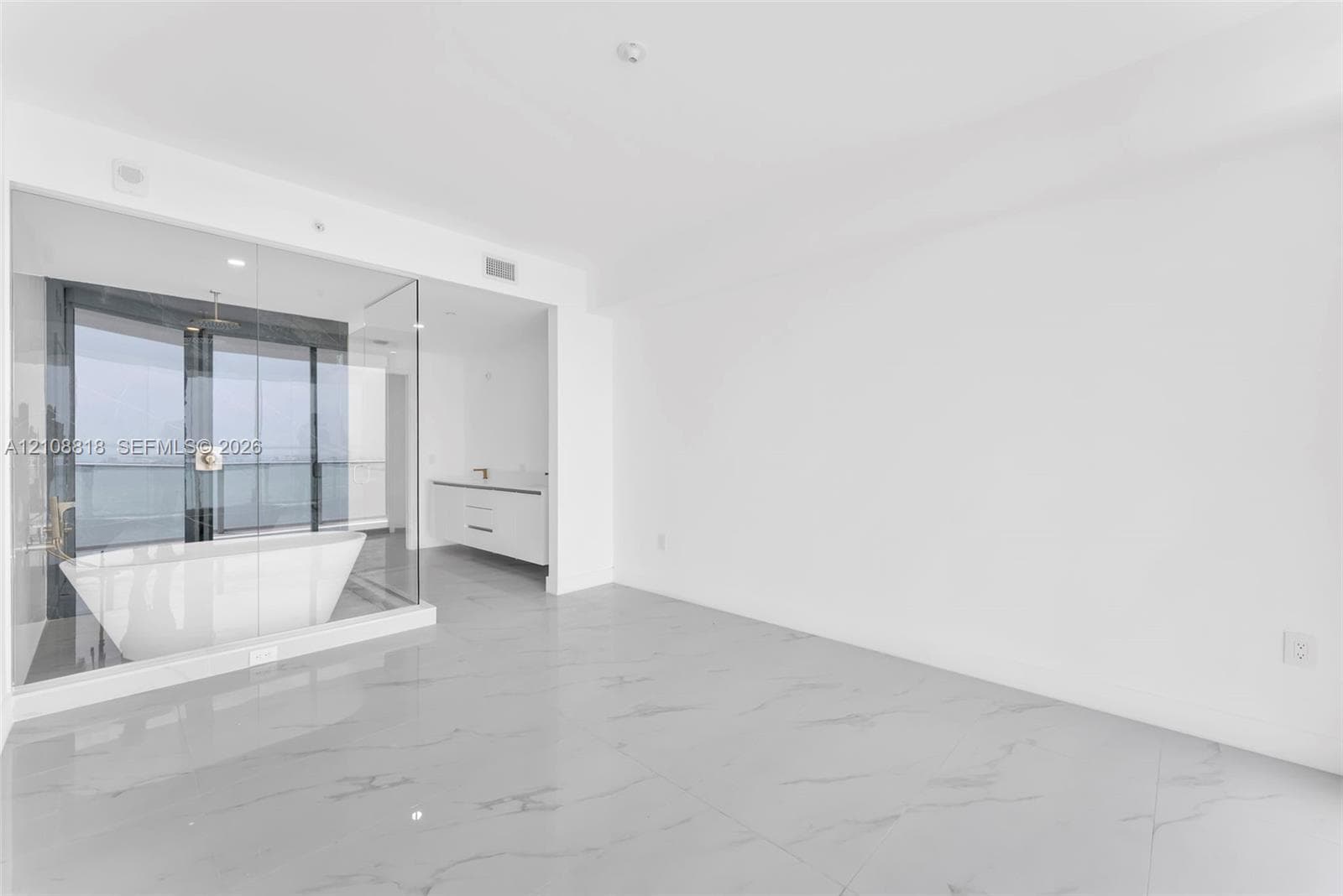 Open-concept space with large-format stone-finish flooring and glass-enclosed soaking tub featuring a wall-mounted faucet and rainfall showerhead, accompanied by a white floating vanity