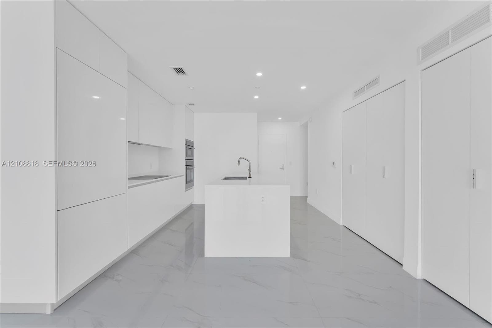 Minimalist kitchen featuring high-gloss cabinetry, a central island with a built-in sink, integrated double wall ovens, recessed ceiling lighting, and large-format polished flooring