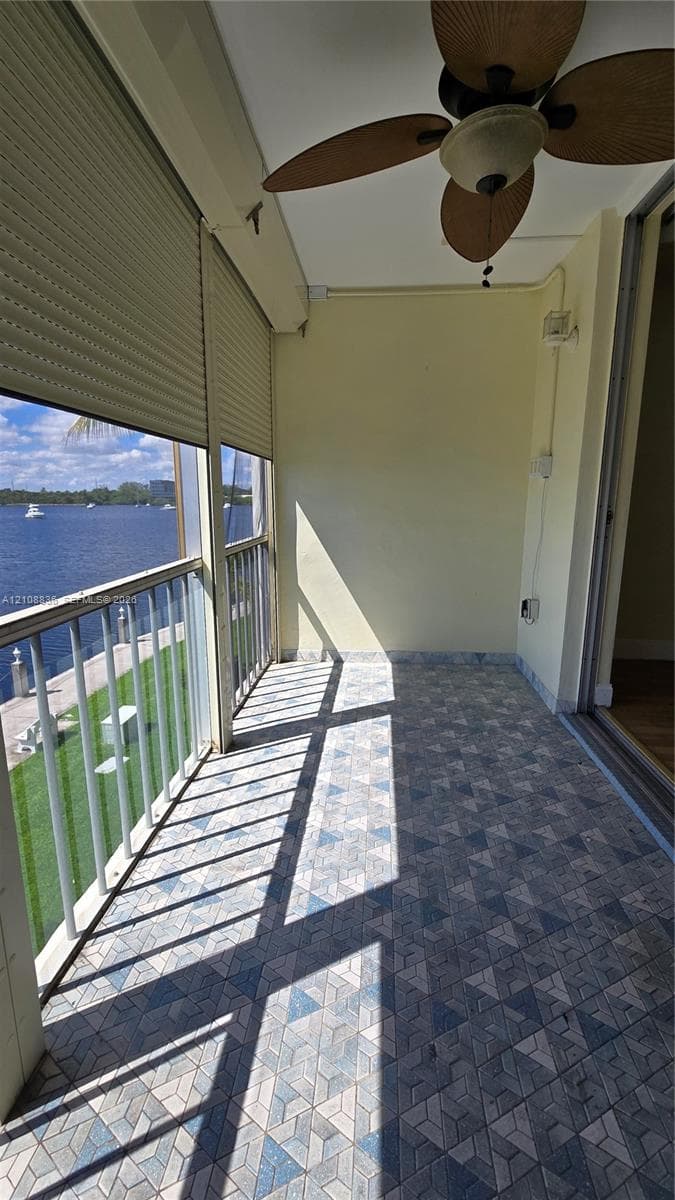 Covered balcony featuring patterned tile flooring, a ceiling-mounted paddle fan, integrated roll-down security shutters, and metal railing overlooking the waterway
