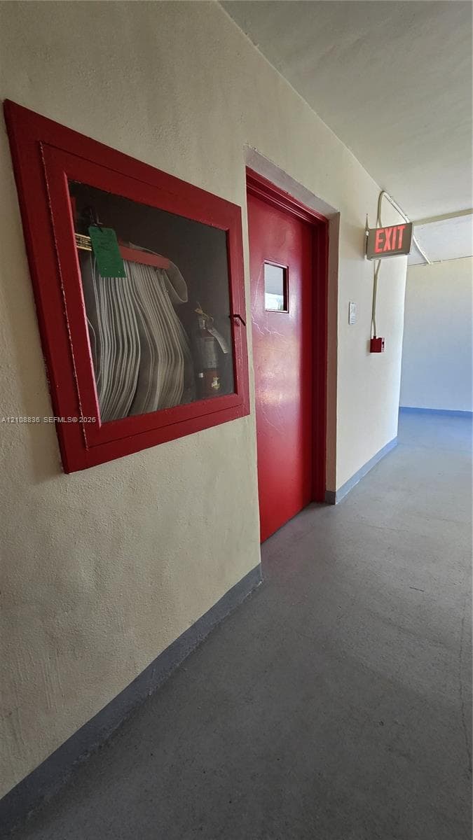 Hallway featuring a painted metal door, wall-mounted fire safety cabinet with glass panel, and illuminated exit signage