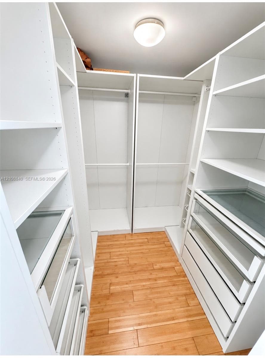 Walk-in closet featuring custom white built-in shelving, double hanging rods, integrated drawers with glass-front detailing, and wood-finish flooring