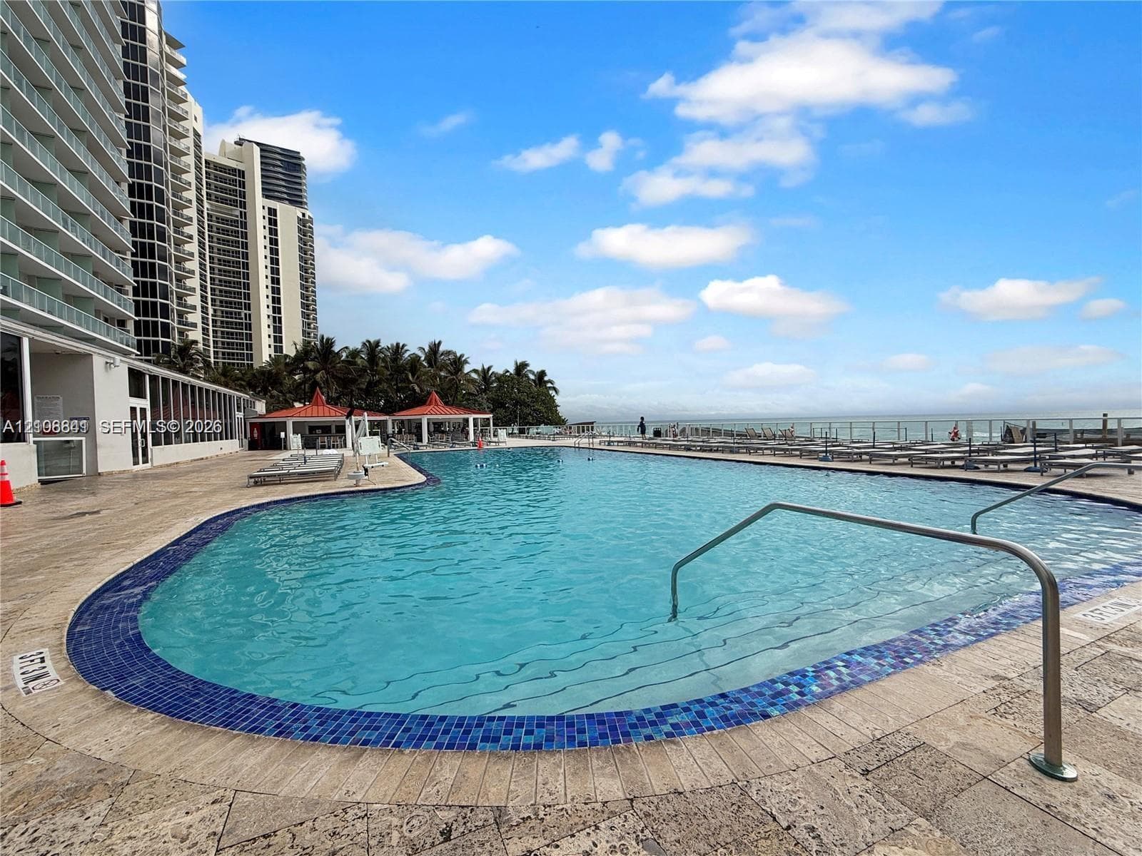 Curved swimming pool featuring blue tile coping, stainless steel handrails, and a natural stone deck